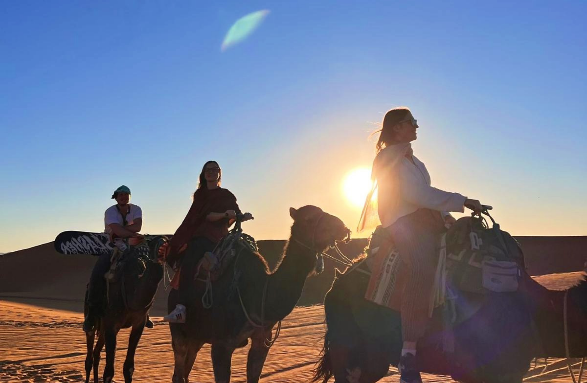 three people atop three camels with sand dunes in the back on a blue sky sunny day